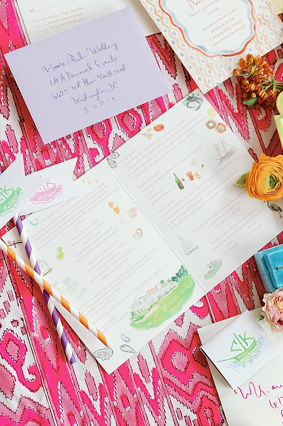 Invitation suite wedding invitation flat lay with calligraphy envelope, illustrated cards, ribbon, and ranunculus on pink patterned textile background