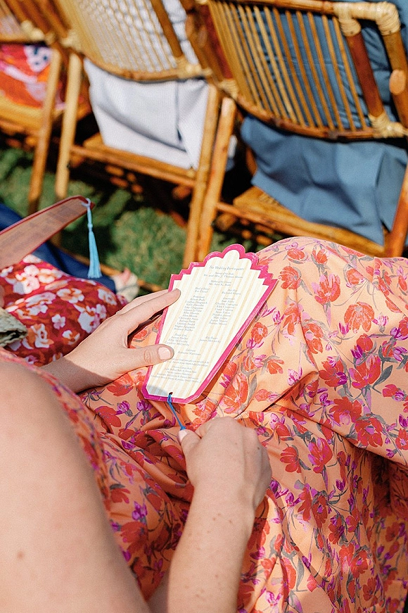 Wedding ceremony program with tassel and scalloped edge held by a guest in a floral dress on a grass lawn beside rattan chairs