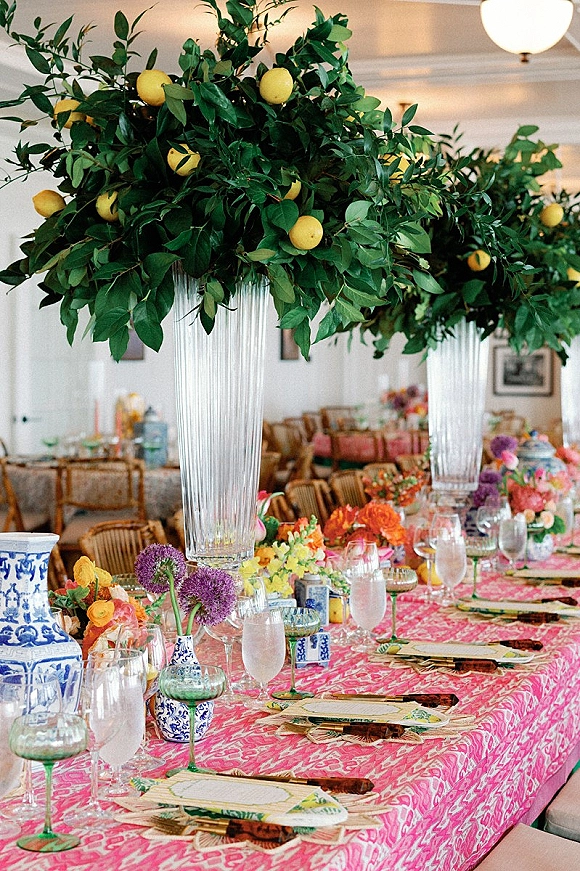 Reception tablescape with pink patterned tablecloth, lush greenery runner, lemons, tall glass vases and colorful florals in an indoor room with framed art