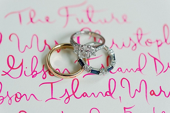 Wedding rings in a wedding ring flat lay with diamond engagement ring, gold band, and sapphire eternity band on pink calligraphy invitation