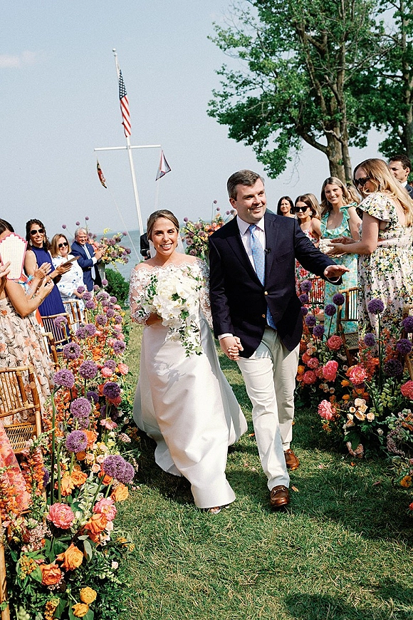 Wedding recessional as bride and groom walk the aisle holding hands, bouquet raised, guests in sunglasses beside florals with ocean view
