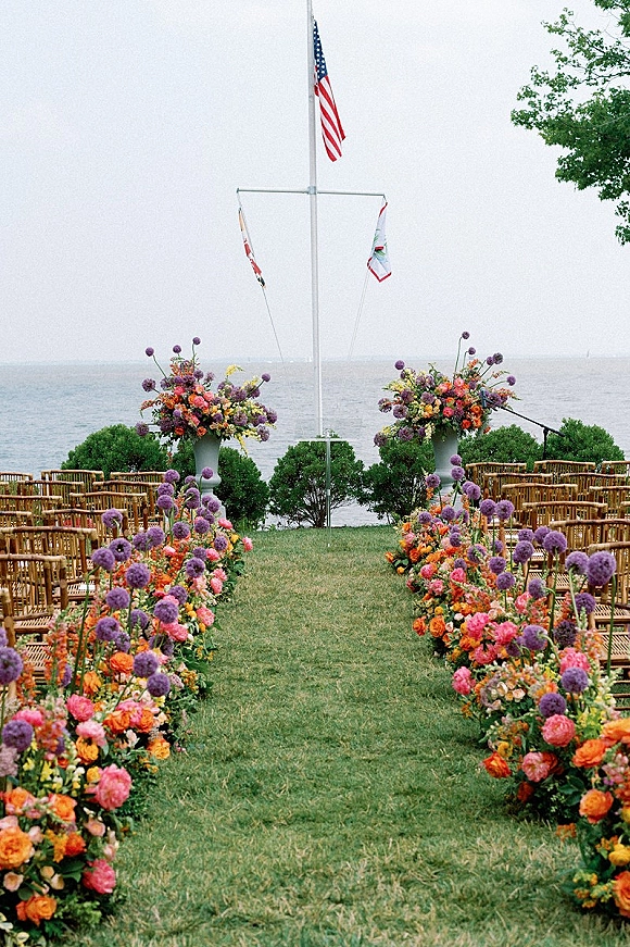 Ceremony aisle decor with floral lined ceremony aisle, purple and orange arrangements along wooden chairs on a waterfront lawn by the ocean