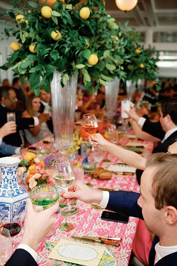Wedding reception toast with wedding toast glasses raised above a pink patterned tablecloth, tall floral centerpiece and hanging globe light indoors