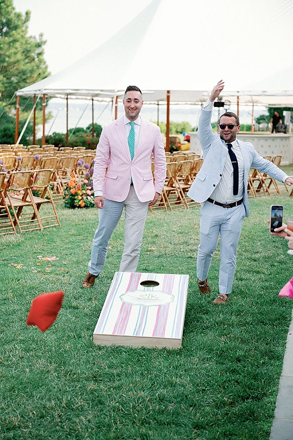 Wedding lawn games with groomsmen playing wedding cornhole on a grassy lawn, pastel suits and sunglasses near ceremony chairs and water view