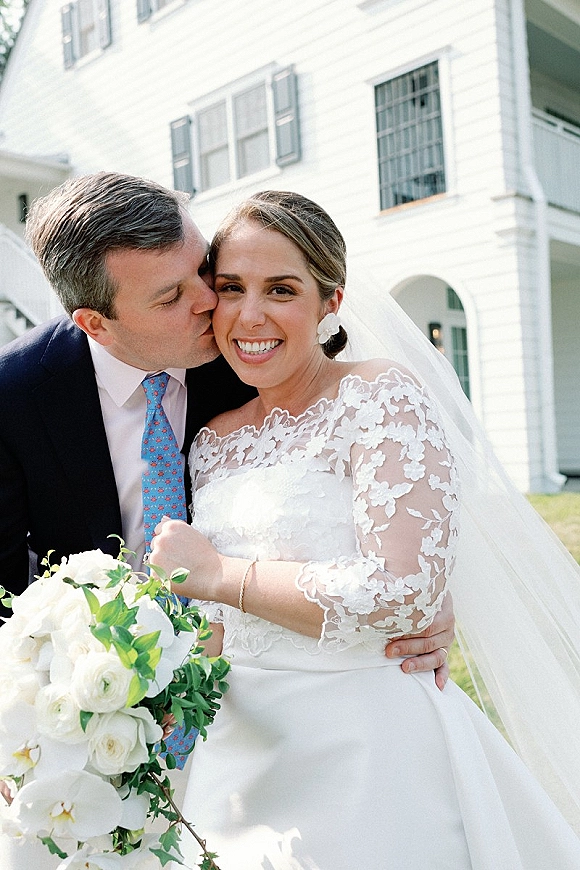 Wedding couple portrait of groom kissing bride’s cheek as she smiles, holding a white rose bouquet, in front of a white house porch