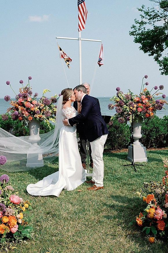 Wedding kiss at an outdoor wedding ceremony, bride in lace gown and long veil kissing groom by waterfront lawn with floral urns