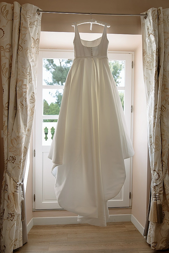 Wedding dress hanging on a hanger before window curtains, softly backlit with curtain tiebacks and a balcony railing beyond