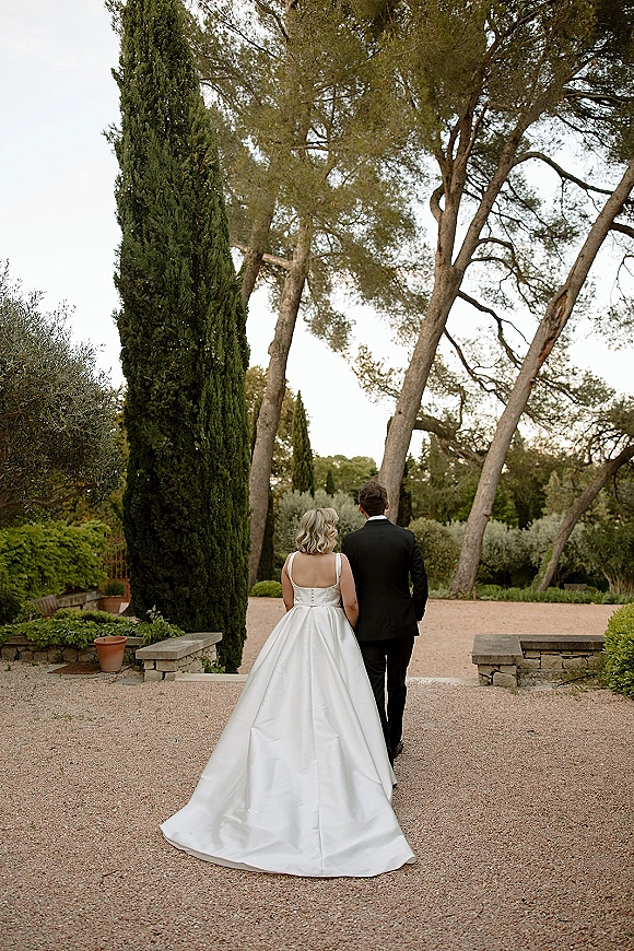 Couple portrait of bride and groom walking away, her dress train trailing on a gravel garden path lined with tall trees