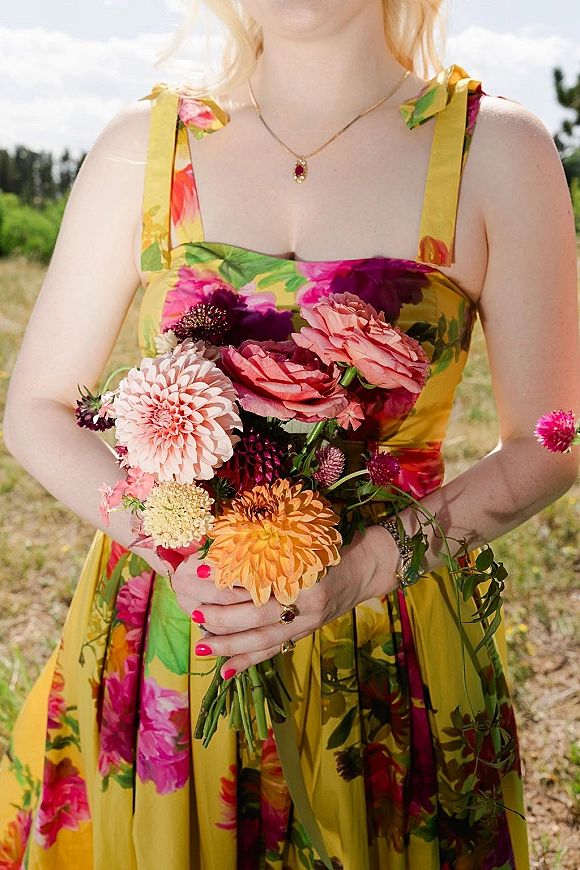Bridesmaid bouquet of colorful dahlias and roses with greenery stems, held against a floral print dress in an outdoor field with trees