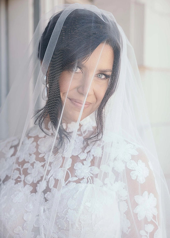 Bridal portrait of a bride with veil draped over her face, showing lace wedding dress and pearl earrings in soft window light indoors
