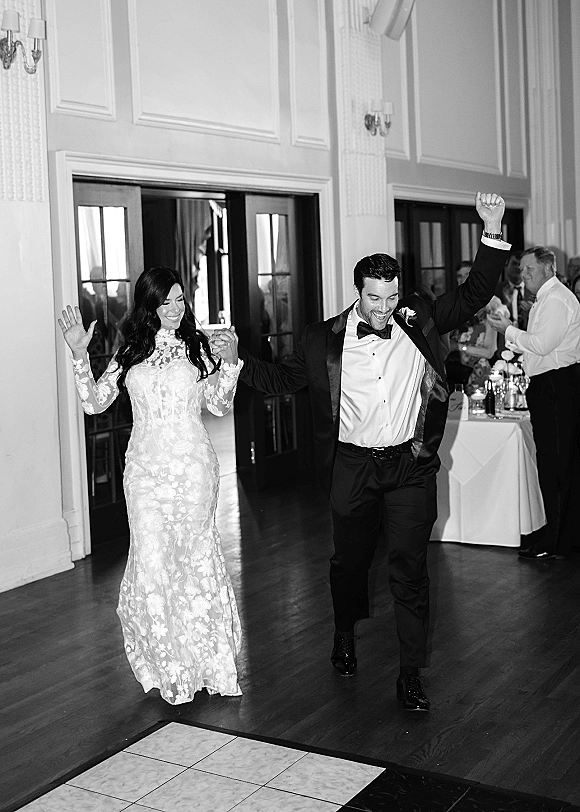 Reception entrance as bride and groom hold hands, her long-sleeve lace gown and his tuxedo, entering a ballroom with cheering guests