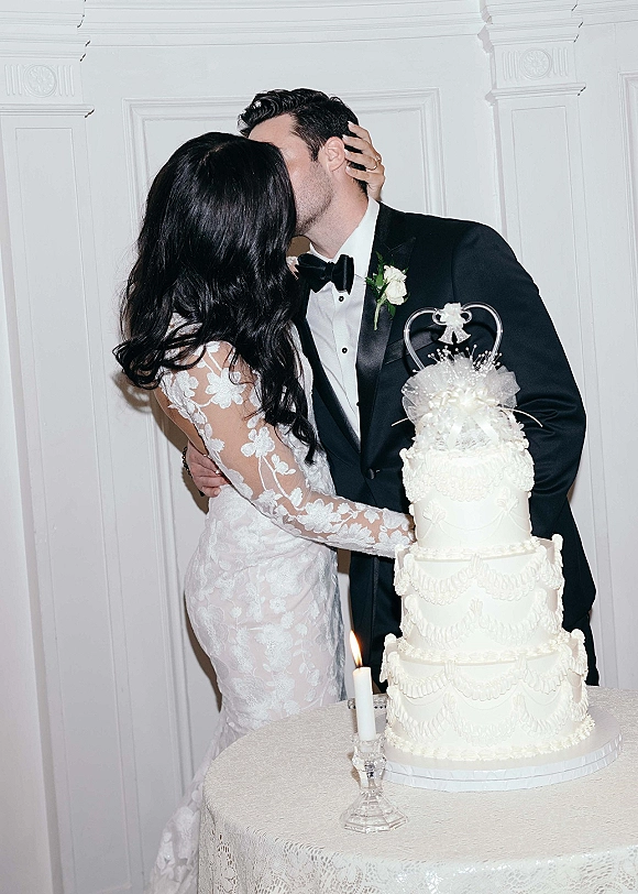Wedding cake cutting with bride and groom kissing beside a classic white three-tier cake on a lace table with a taper candle indoors