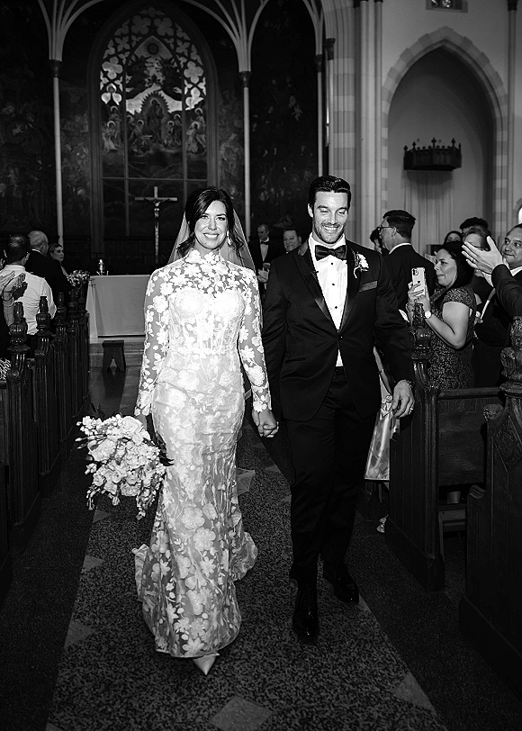 Wedding recessional as bride and groom walk hand in hand down a church aisle, her lace long-sleeve gown and bouquet as guests cheer beneath gothic arches