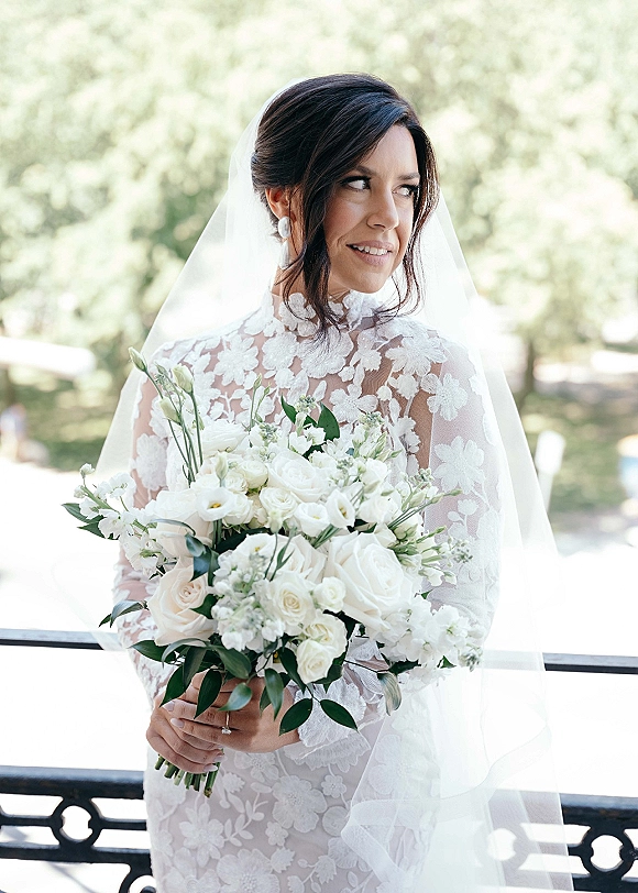 Bridal portrait of a bride holding bouquet of white roses in a lace long-sleeve dress and cathedral veil, standing in a sunlit garden.