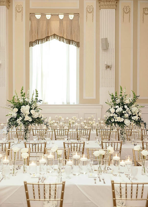 Reception tablescape with white wedding reception tables, tall floral and greenery centerpieces, candles, and gold Chiavari chairs in a classic ballroom