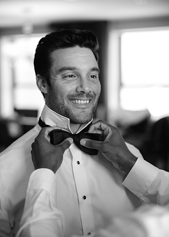 Groom getting ready, adjusting a black bow tie on his tuxedo shirt with stud accents in soft window light, blurred room behind