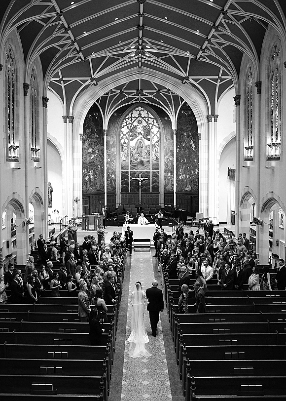 Wedding processional with bride walking down aisle in a long veil and train between church pews toward a cross-lit altar under chandeliers