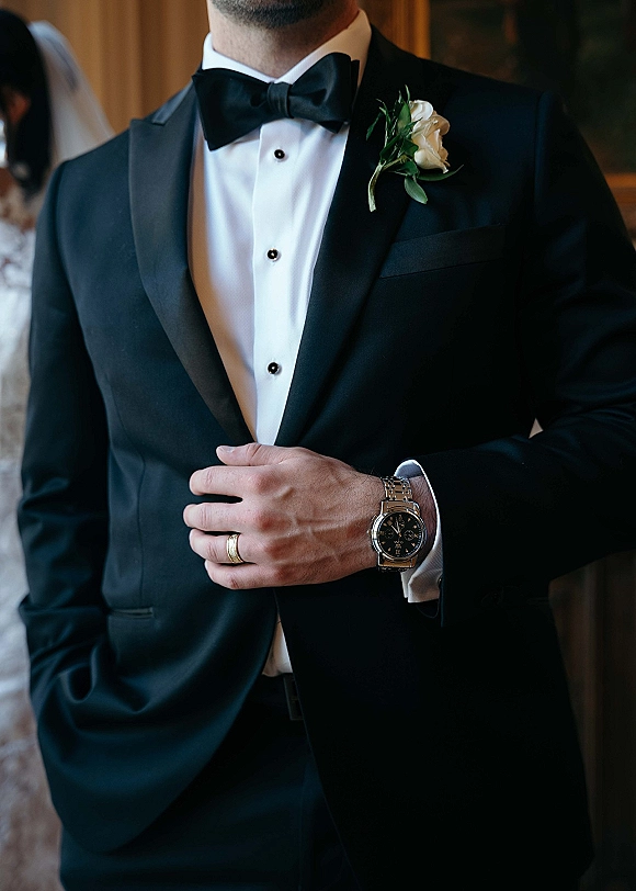 Groom portrait in a black tuxedo with satin lapel and bow tie, wearing a white rose boutonniere, bride softly blurred behind indoors