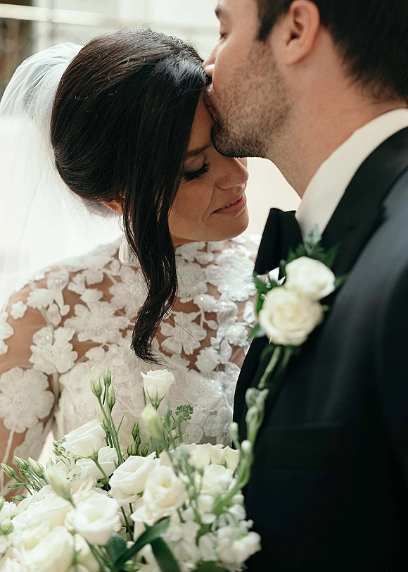 Wedding couple portrait of groom kissing bride’s forehead as she holds a white rose bouquet, lace dress and veil glowing in window light