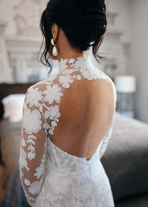 Wedding dress with illusion lace high neck and open back, styled with bridal updo and pearl drops beside a bedroom bed and lamp