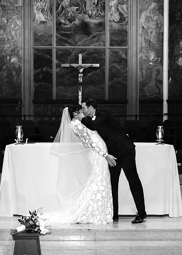 Wedding kiss at the altar as bride in long-sleeve lace gown and veil leans into groom in tuxedo, crucifix and candles behind