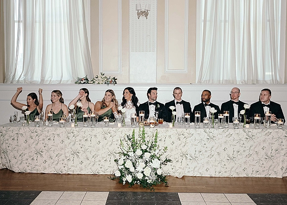 Wedding head table with bride and bridal party seated amid candles, white roses, and greenery, set against a white paneled wall backdrop