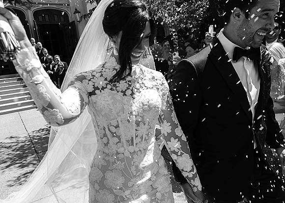 Wedding recessional as bride in long sleeve lace dress and veil holds hands with groom in tuxedo amid confetti on church steps
