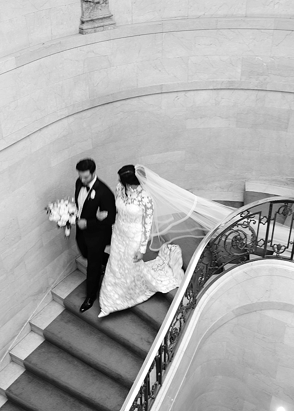 Wedding couple portrait of bride and groom on staircase, arm in arm, her lace gown and long veil flowing beside a bouquet of white flowers.