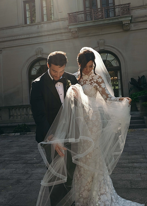 Couple portrait of bride and groom portrait with a flowing wedding veil on stone steps before an arched-window facade and courtyard plants