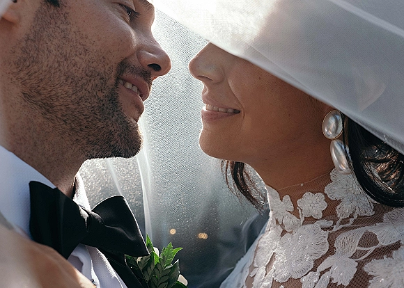 Wedding couple portrait of bride and groom under veil, forehead to forehead in soft sunlight bokeh, her lace dress and pearl earrings visible
