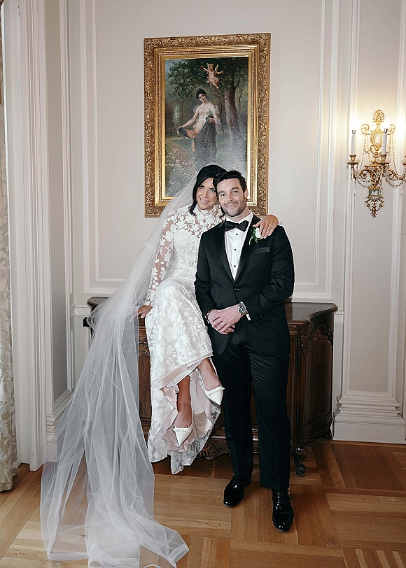 Couple portrait in an indoor wedding portrait, bride in lace sleeves and long veil seated on a sideboard beside groom in tuxedo under an ornate gold frame