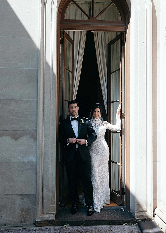 Couple portrait of bride in a high neck lace wedding dress and veil with groom in tuxedo, framed by an arched stone doorway