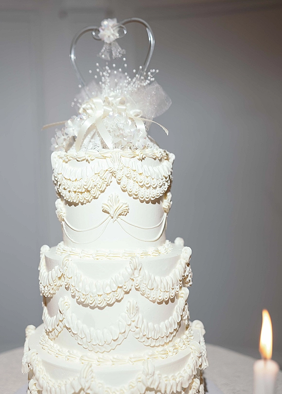 Wedding cake with piped buttercream swags on a white tiered wedding cake, finished with a heart topper and ribbon bow on a table