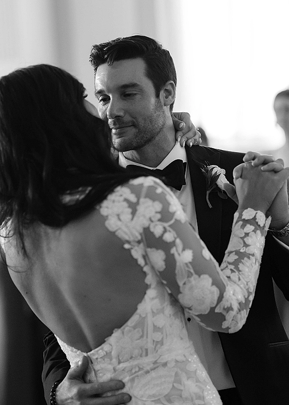 First dance close up of bride in a low-back lace sleeve wedding dress embracing groom in tuxedo, bright window light and blurred guests behind