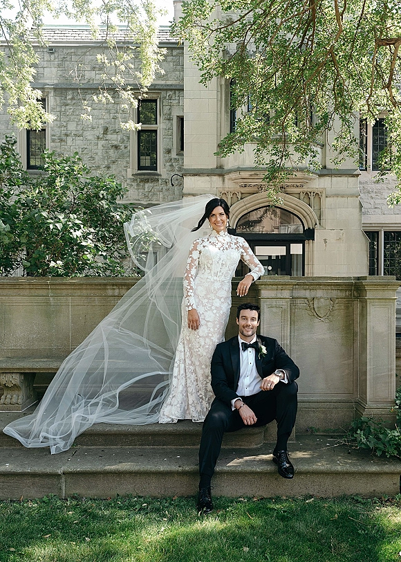Couple portrait of bride in lace dress and long veil leaning on groom in tuxedo seated on stone steps by an arched doorway