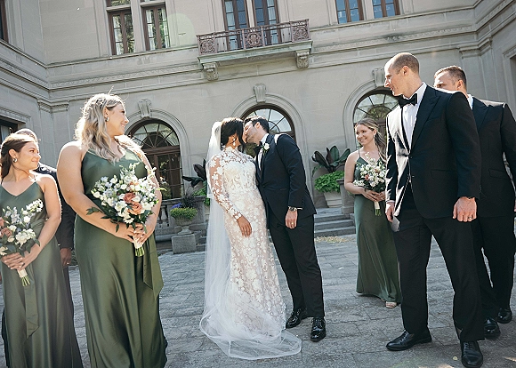 Wedding party portrait as the bride and groom kiss, surrounded by bridesmaids and groomsmen in black tuxedos on stone mansion steps.