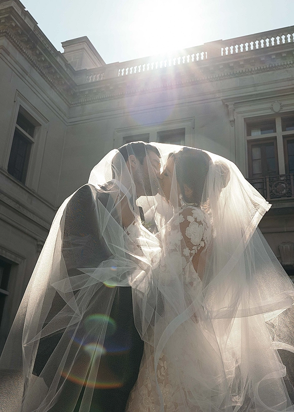 Wedding kiss portrait of bride and groom under a veil, lace dress and boutonniere glowing with sun flare by a stone building balcony