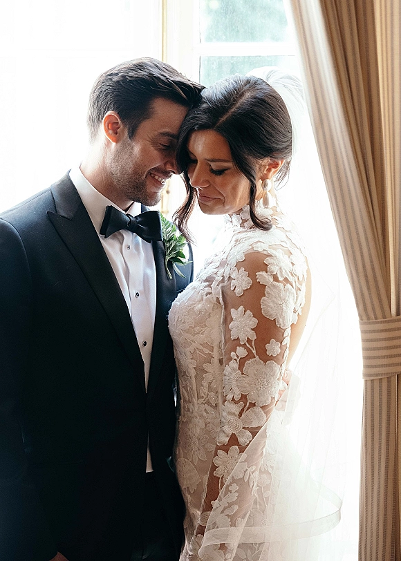 Couple portrait of bride and groom portrait touching foreheads by a window, groom in black tuxedo and bride in lace sleeves with veil