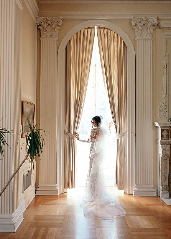 Bridal portrait of a bride by window in a lace long sleeve wedding dress, holding curtains as her veil trails on hardwood floors indoors
