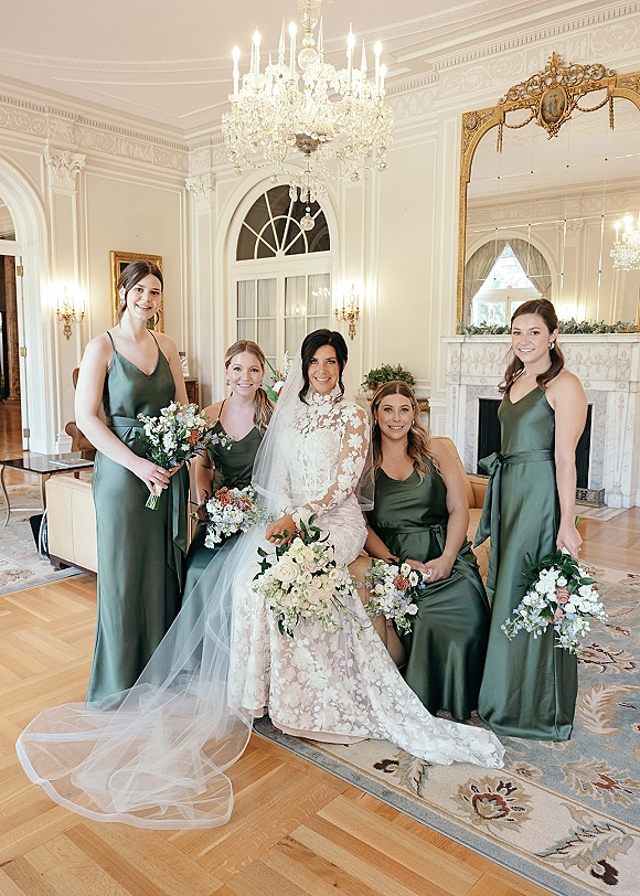 Bride with bridesmaids posing in green bridesmaid dresses, holding bouquets in an elegant room with arched windows and crystal chandelier