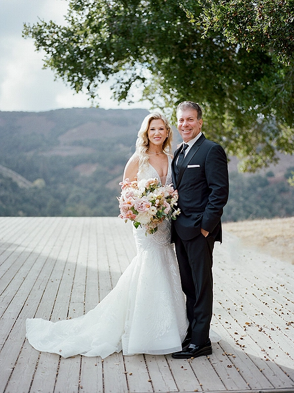 Couple portrait on a wooden deck with mountain views, bride in a lace mermaid wedding dress holding a pastel bouquet beside groom in tuxedo