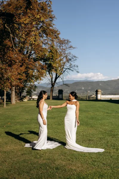 Couple portrait of two brides holding hands and smiling in long train wedding dresses on a lawn with autumn trees and mountains behind