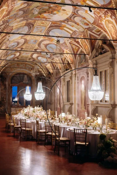 Reception tablescape with round wedding tables, white linens, low floral centerpieces and taper candles beneath crystal chandeliers in a frescoed hall