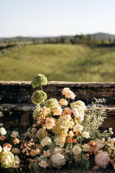 Wedding ceremony florals in a ground arrangement with hydrangeas and roses, soft greenery accents, set against a stone wall and hillside sky