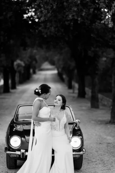 Couple portrait of two brides in wedding dresses holding champagne flutes beside a vintage car on a tree-lined gravel driveway