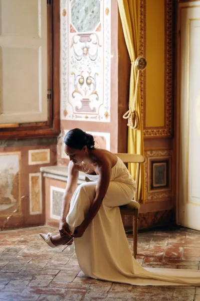 Bridal portrait of a bride putting on shoes, tying pearl wedding heels while seated in a strapless gown in an ornate room by a large window