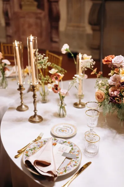 Reception tablescape with a wedding table centerpiece of peach and blush florals, taper candles, gold flatware, and crystal glassware indoors