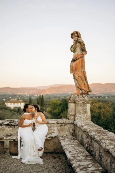 Couple portrait of a lesbian wedding couple kissing in wedding dresses, holding champagne flutes on a stone terrace by a villa statue