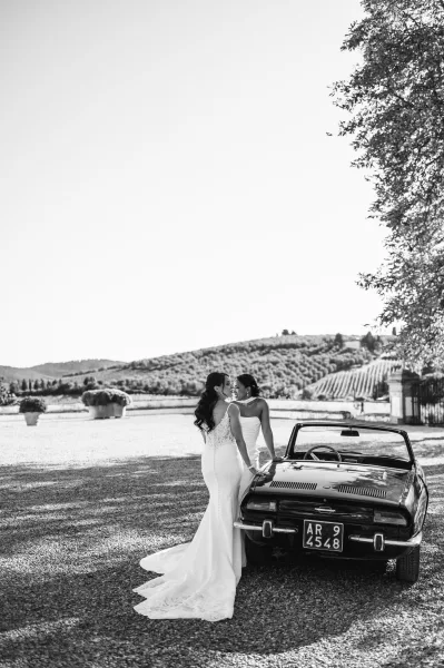 Couple portrait of two brides by a vintage convertible car, one in a lace backless gown and veil, on a gravel vineyard drive
