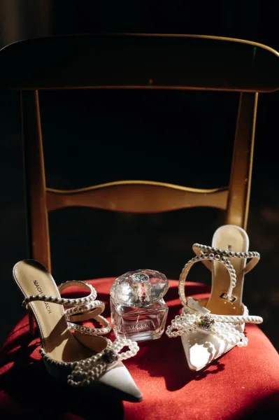 Bridal shoes, pearl wedding heels with rhinestone buckle and pointed toe, styled beside perfume on a red velvet chair in dark interior